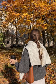 a woman standing in front of a tree with leaves on the ground and holding a cup