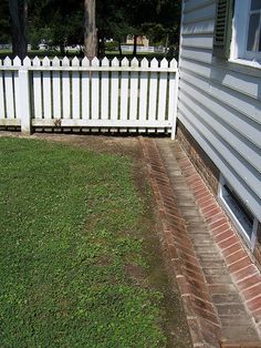 a white picket fence in front of a house with grass on the ground and brick walkway