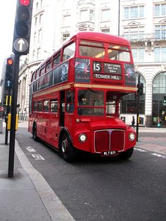 a red double decker bus driving down a street next to a traffic light and tall buildings