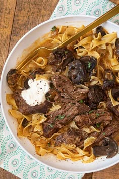 a white bowl filled with pasta and meat on top of a wooden table next to a spoon