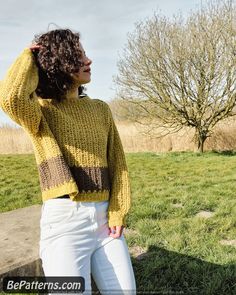 a woman standing in front of a tree wearing a yellow sweater and white pants with her hands on her head