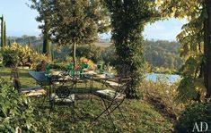 an outdoor dining table and chairs in the grass near trees, water and plants on either side