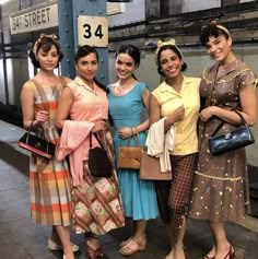 four women standing next to each other in front of a train station sign and holding purses