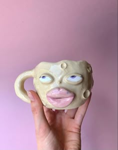 a person holding up a ceramic mask with blue eyes and pink cheeks on a pink background