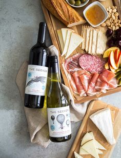 two bottles of wine, cheese and meats on a cutting board with bread slices