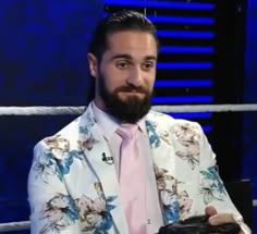 a man with a beard wearing a white suit and pink tie sitting in a chair