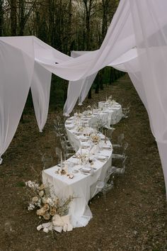 a long table set up with white linens and flowers for a wedding reception in the woods