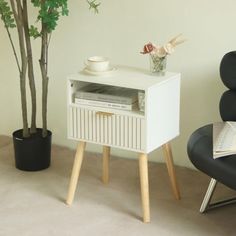 a small white cabinet next to a black chair and a potted tree in a corner