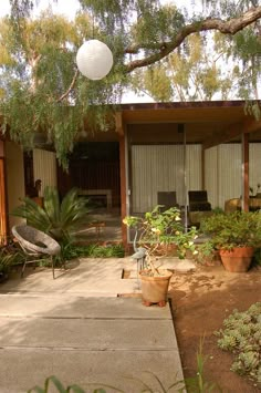 an outdoor patio area with potted plants and chairs under a tree in front of a house