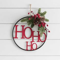 a christmas wreath hanging on the side of a white wall with red letters and pine cones