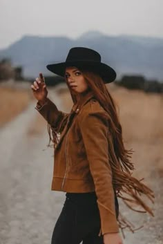 a woman with long hair wearing a brown jacket and black hat walking down a dirt road