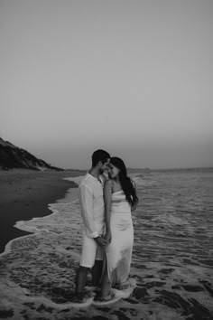 a man and woman standing on top of a sandy beach next to the ocean holding each other