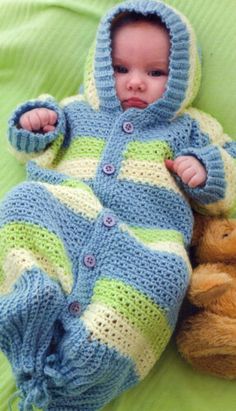 a baby in a blue and green knitted outfit laying next to a teddy bear