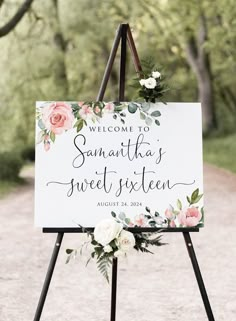 a welcome sign with flowers and greenery is displayed on an easel in the middle of a dirt road