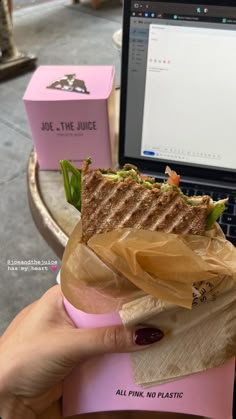 a person holding a sandwich in front of a laptop on a table with pink boxes