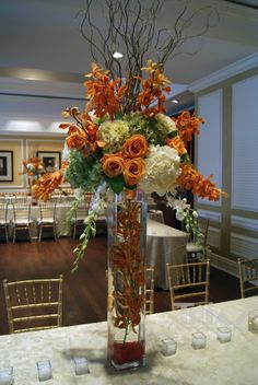 an arrangement of flowers in a tall vase on a table with chairs and tables around it