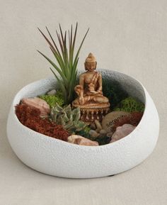 a buddha statue sitting in a bowl filled with rocks and succulents next to a plant