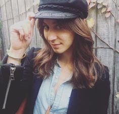 a woman wearing a black hat and jacket standing in front of a fence with her hand on her hip