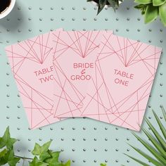 two pink wedding place cards sitting on top of a table next to plants and potted plant