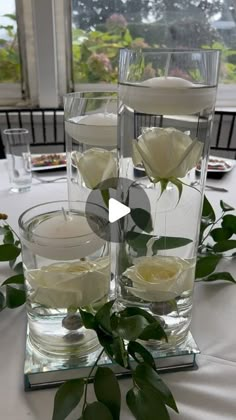 two clear vases filled with white flowers on top of a table covered in greenery