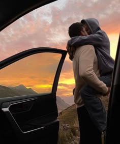 two people standing in the open door of a car with mountains in the background at sunset