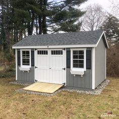 a gray and white shed with two windows on the roof, next to some trees