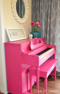 a pink piano in front of a window with curtains and flowers on the table next to it