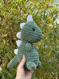 a hand holding a green crocheted stuffed animal in front of some trees and bushes