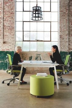 two women sitting at a table talking to each other in an office setting with large windows