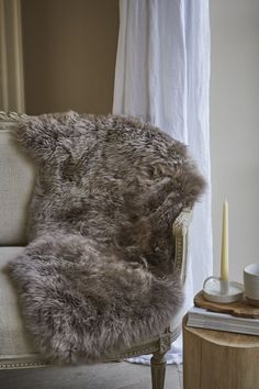 a chair with a fur cushion on it next to a table and candle in front of a window