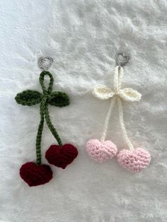 two crocheted cherries hanging from hooks on a white blanket, one is pink and the other is green