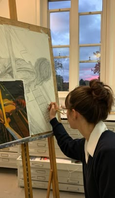 a woman is drawing on an easel in a room with windows and other items