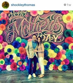 a man and woman standing in front of a flower wall with the words happy mother's day written on it