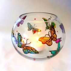 a glass bowl with butterflies painted on it and a lit candle in the middle, sitting on a white surface