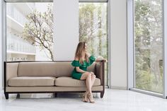 a woman in a green dress sitting on a couch looking out the window at trees