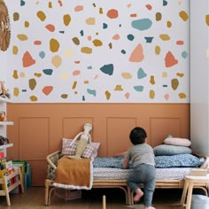 a little boy sitting on top of a bed in front of a wall with colorful shapes