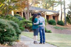 an engaged couple standing on a path in front of a house with trees and bushes