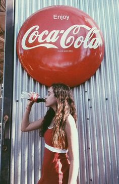 a woman in a red dress drinking from a coca - cola bottle next to a metal wall