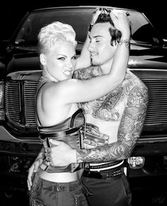 a man and woman hugging each other in front of a car with tattoos on their arms