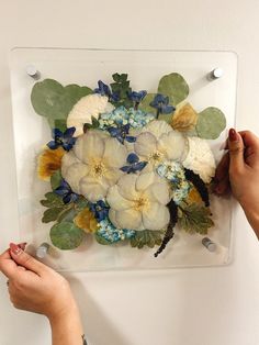 two hands are working on a floral arrangement in a glass frame with acrylic paint