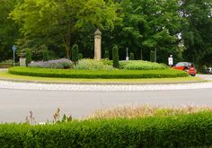 a red car driving down a street next to a lush green park filled with trees