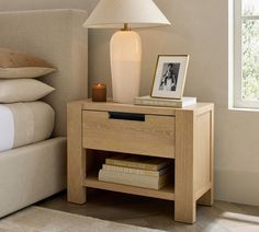 a night stand with a lamp on it next to pillows and a bed in the background