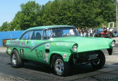 an old green car driving down the road with people standing around it and onlookers watching