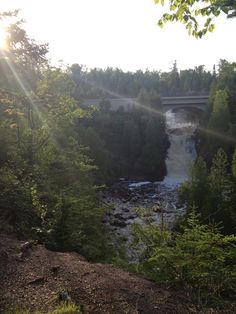 the sun shines brightly in front of a bridge over a river with rapids and trees