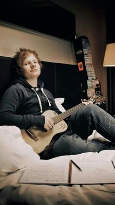 a young man sitting on top of a bed holding an acoustic guitar and looking up at the sky