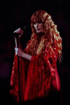 a woman with long red hair holding a microphone in her right hand and wearing a red dress