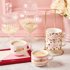 two cups of coffee sit on a table next to a pink vase with white flowers