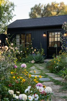 a small black house with lots of flowers in the front yard and walkway leading to it