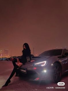 a woman sitting on the hood of a sports car in front of a city at night