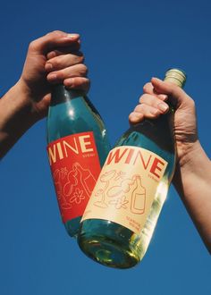 two people holding up wine bottles in the air with blue sky behind them and one person reaching for it